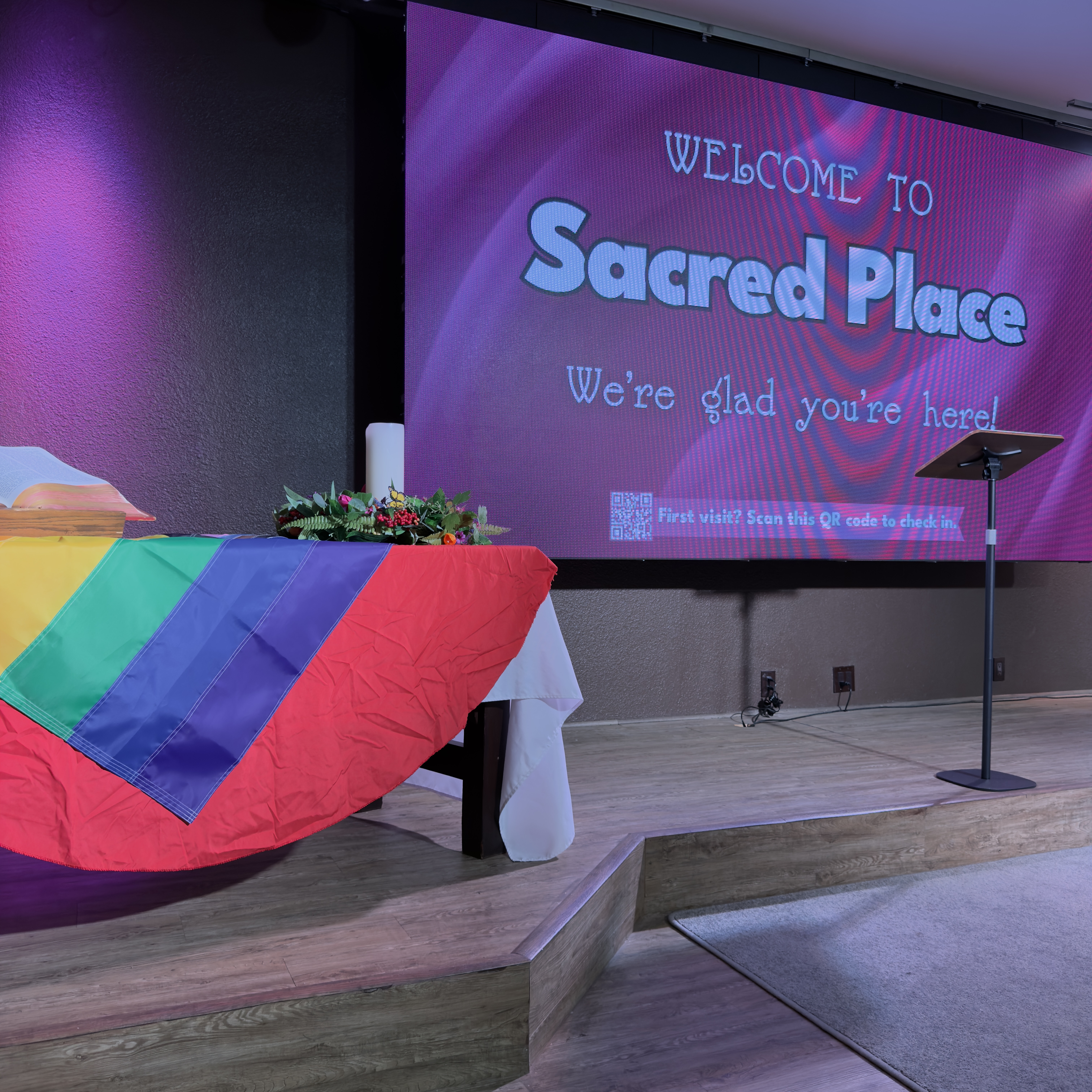A rainbow Pride flag draped over the communion table at Sacred Place, beneath a “Welcome to Sacred Place” banner.